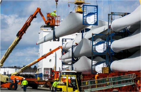 Dock workers and crane operators load large wind turbine blades onto a cargo ship. Multiple white blades are stacked in blue transport frames, and workers in high-visibility gear operate machinery to secure and guide the heavy equipment. A clear blue sky and industrial shipping equipment fill the background.