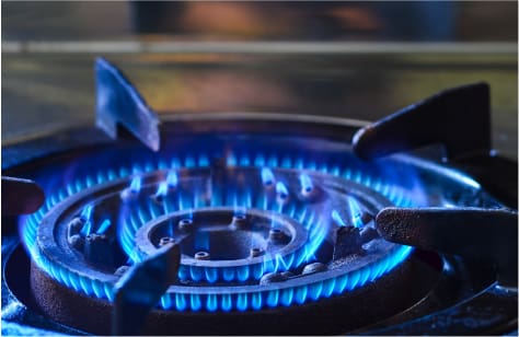 Close-up of a lit gas stove burner emitting an even, bright blue flame. The cast iron grate surrounds the burner, and the background shows a blurred kitchen surface, highlighting clean and efficient natural gas combustion.