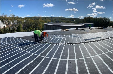 Workers in safety gear install curved rows of solar panels on a flat commercial rooftop under a sunny blue sky. The surrounding area includes forested hills and industrial buildings in the background, indicating a renewable energy project in progress.