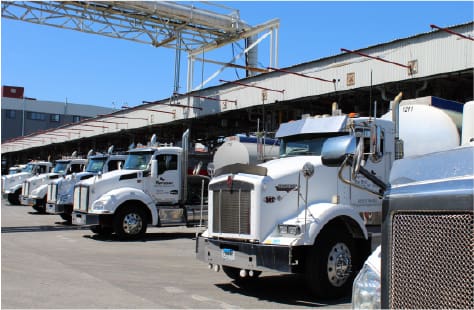 Row of white Sprague-branded fuel delivery trucks parked outside a large terminal building under a clear blue sky, showcasing fleet readiness and industrial infrastructure.