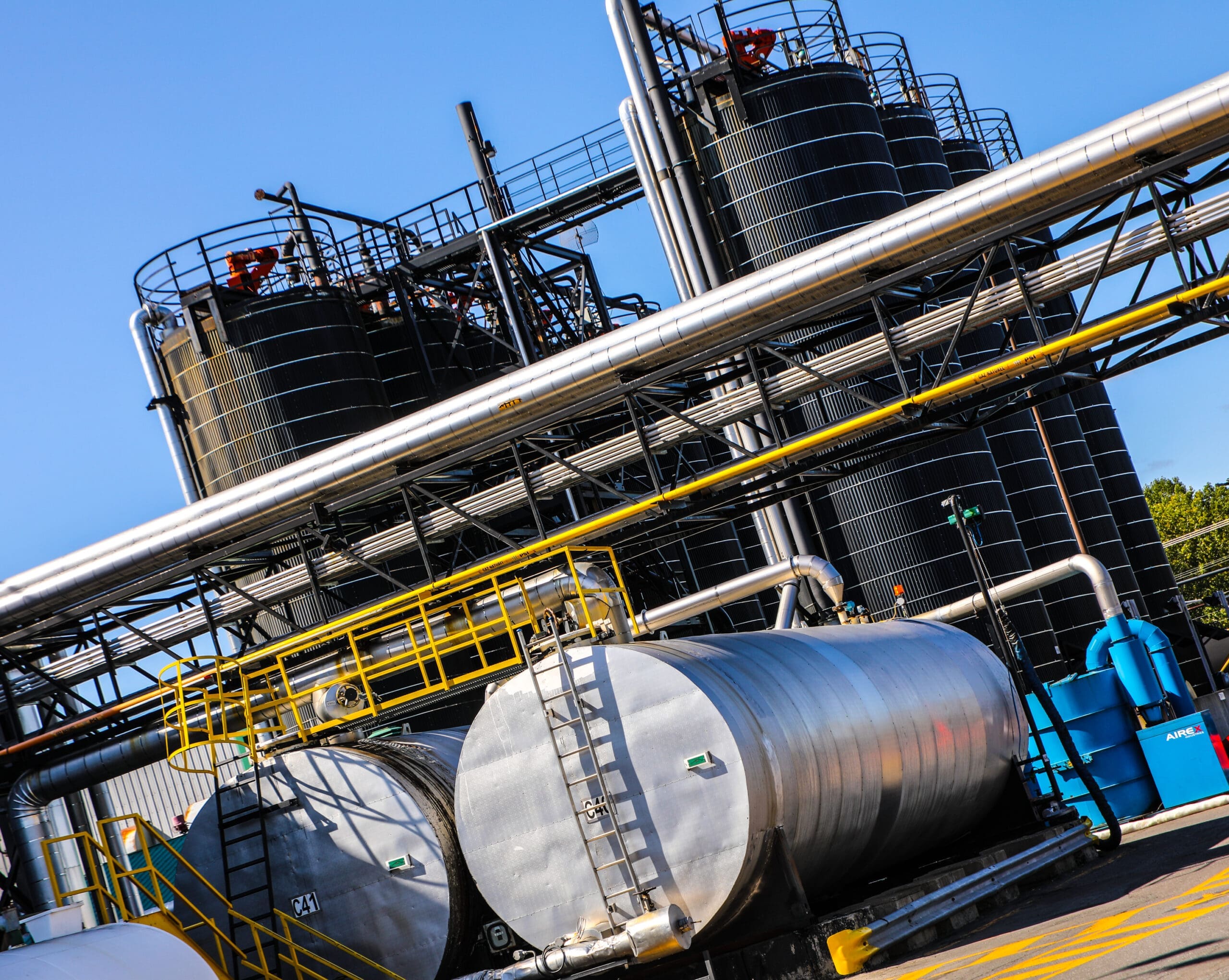 Industrial facility with large cylindrical metal storage tanks and multiple interconnected pipes. The image features silver horizontal tanks in the foreground and tall black vertical tanks in the background, along with walkways, ladders, and safety railings, all under a clear blue sky.