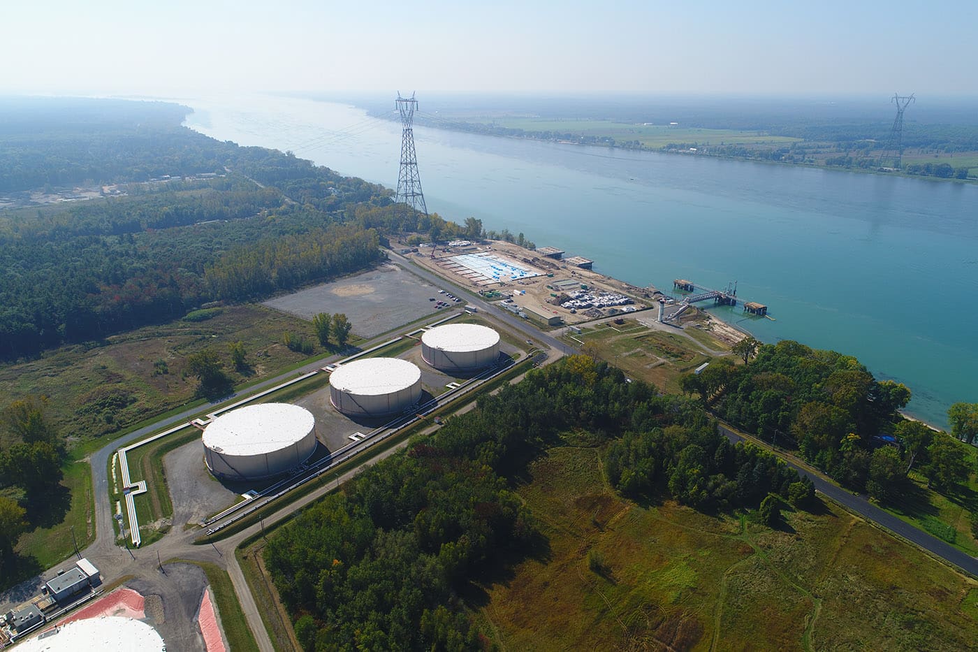 Aerial view of a riverside industrial site featuring three large white cylindrical storage tanks, surrounded by trees and open land. The facility is located along a wide river, with additional infrastructure and utility towers extending along the shoreline.