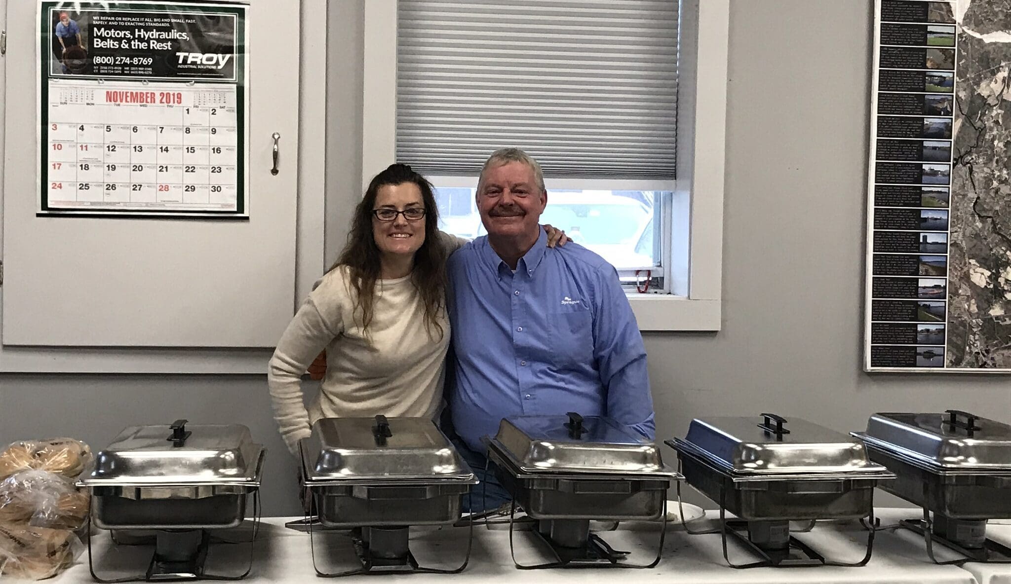 Two smiling people standing behind a row of chafing dishes set up for a meal service, with arms around each other. A calendar on the wall shows November 2019, indicating a past office or community event.