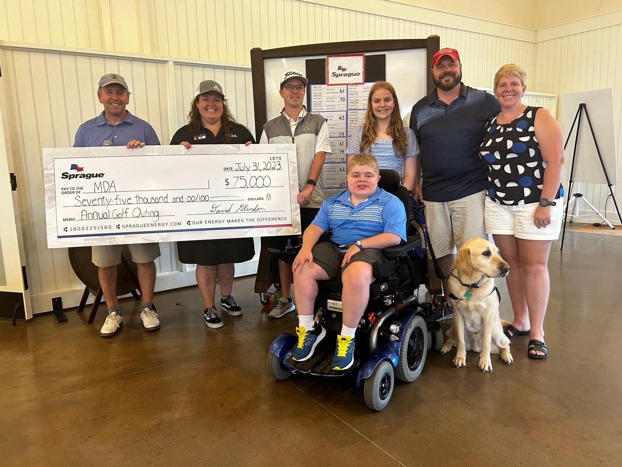 Group of people smiling indoors, including a young person in a wheelchair with a service dog, posing with a large ceremonial check for a charitable donation. A tournament scoreboard is displayed in the background.