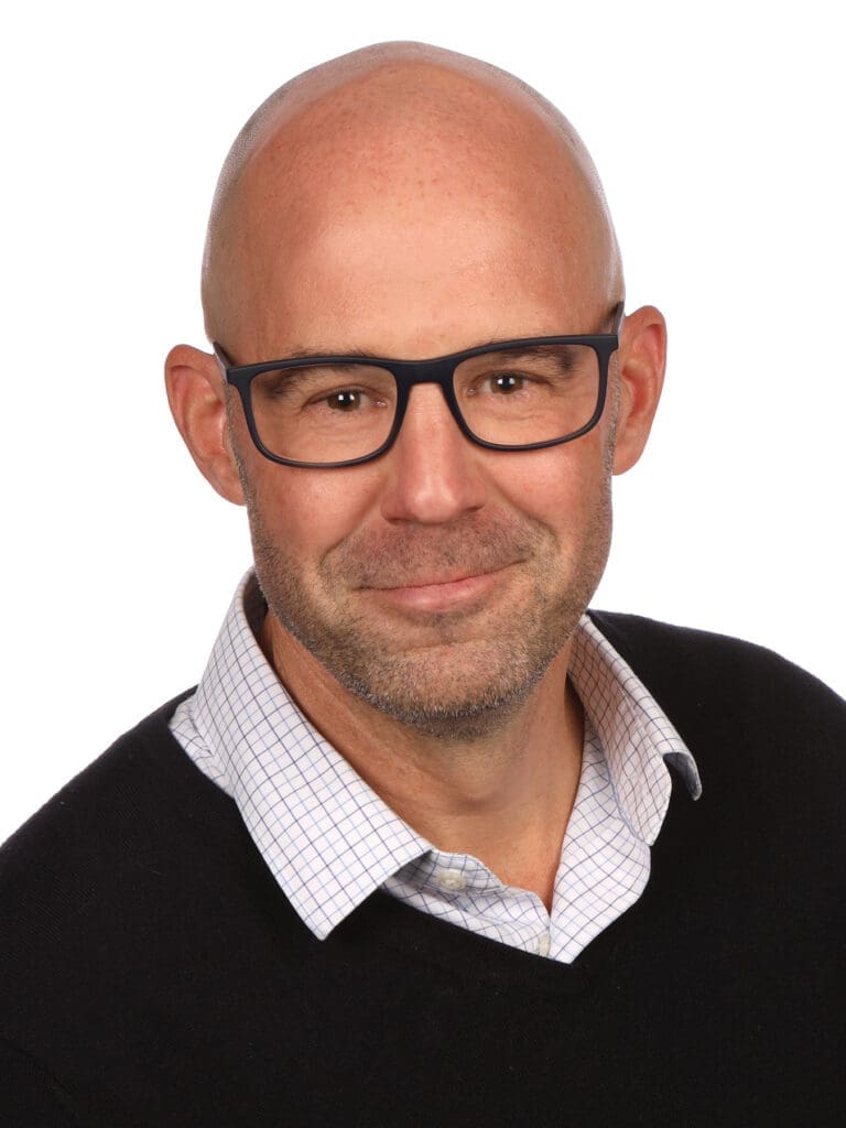 Headshot of Marc Lemay, wearing glasses, a collared shirt, and a dark sweater, smiling in front of a white background.