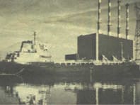 Sepia-toned photo of a cargo ship docked at an industrial port with multiple tall smokestacks in the background.