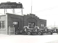 Black and white photo of a vintage Delaney’s fuel company building with four branded delivery trucks parked out front.