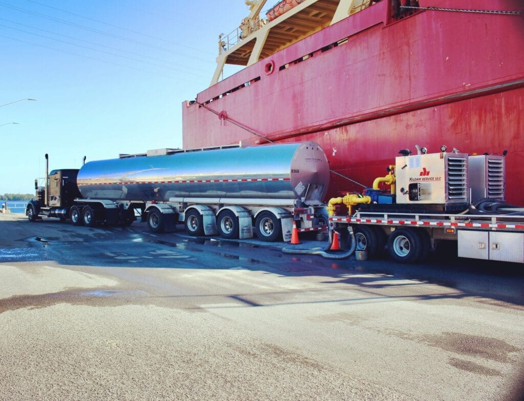 A fuel tanker truck is connected to a large red cargo ship for fueling operations at a port. Equipment and hoses are set up on the ground, with safety cones marking the area.