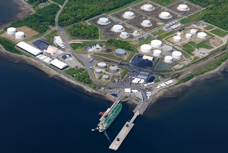 Aerial view of an industrial waterfront facility with multiple storage tanks, cargo areas, and a green ship docked at a pier, surrounded by trees and water.