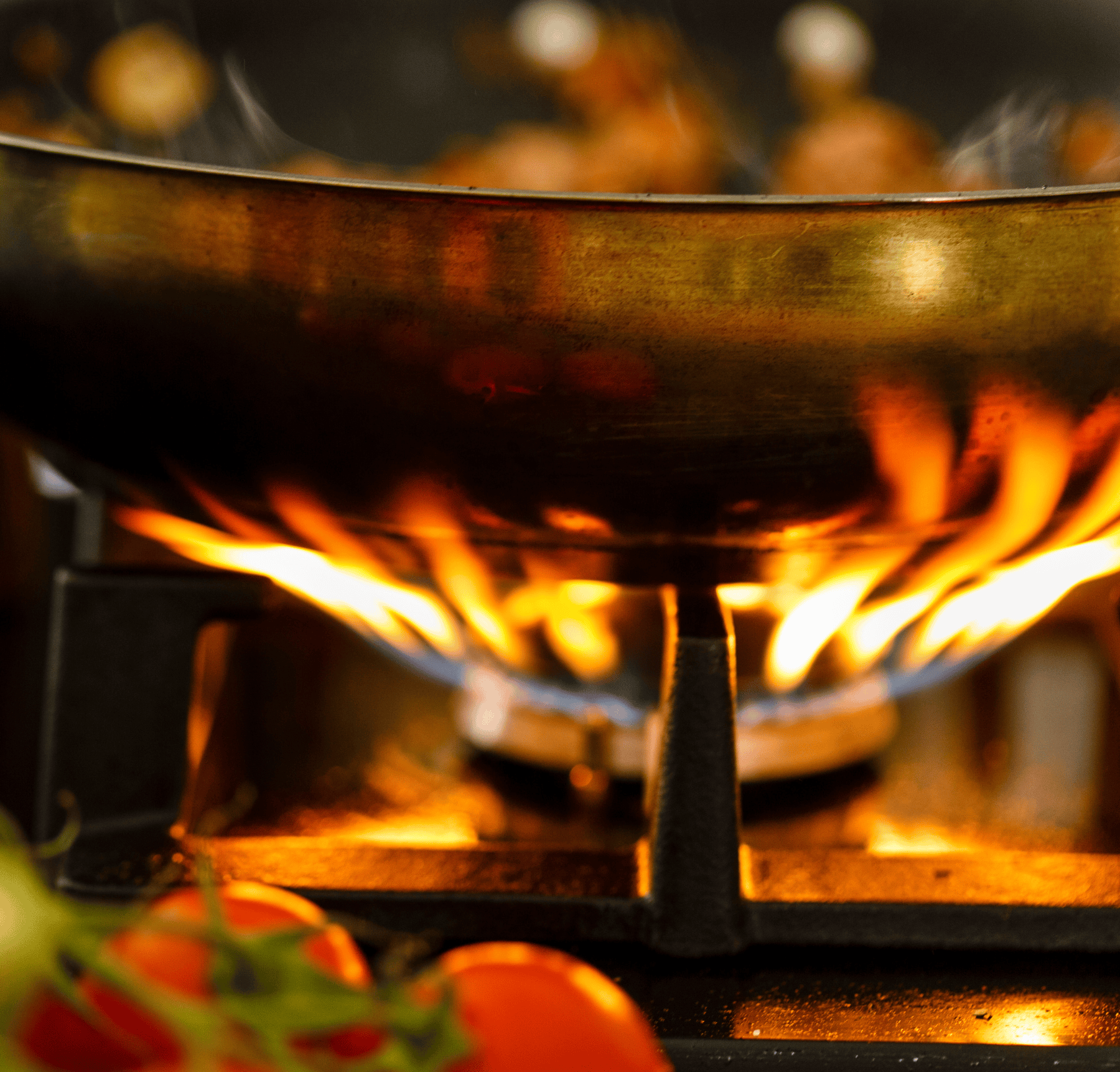 Close-up of a pan over an open gas flame on a commercial stove, with cherry tomatoes in the foreground, emphasizing natural gas cooking in a professional kitchen.