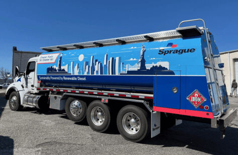 Sprague-branded fuel delivery truck with a vibrant blue wrap featuring a city skyline, wind turbines, and the Statue of Liberty, labeled "Sustainably Powered by Renewable Diesel" and "Cleaner Fuels for Cleaner Air."