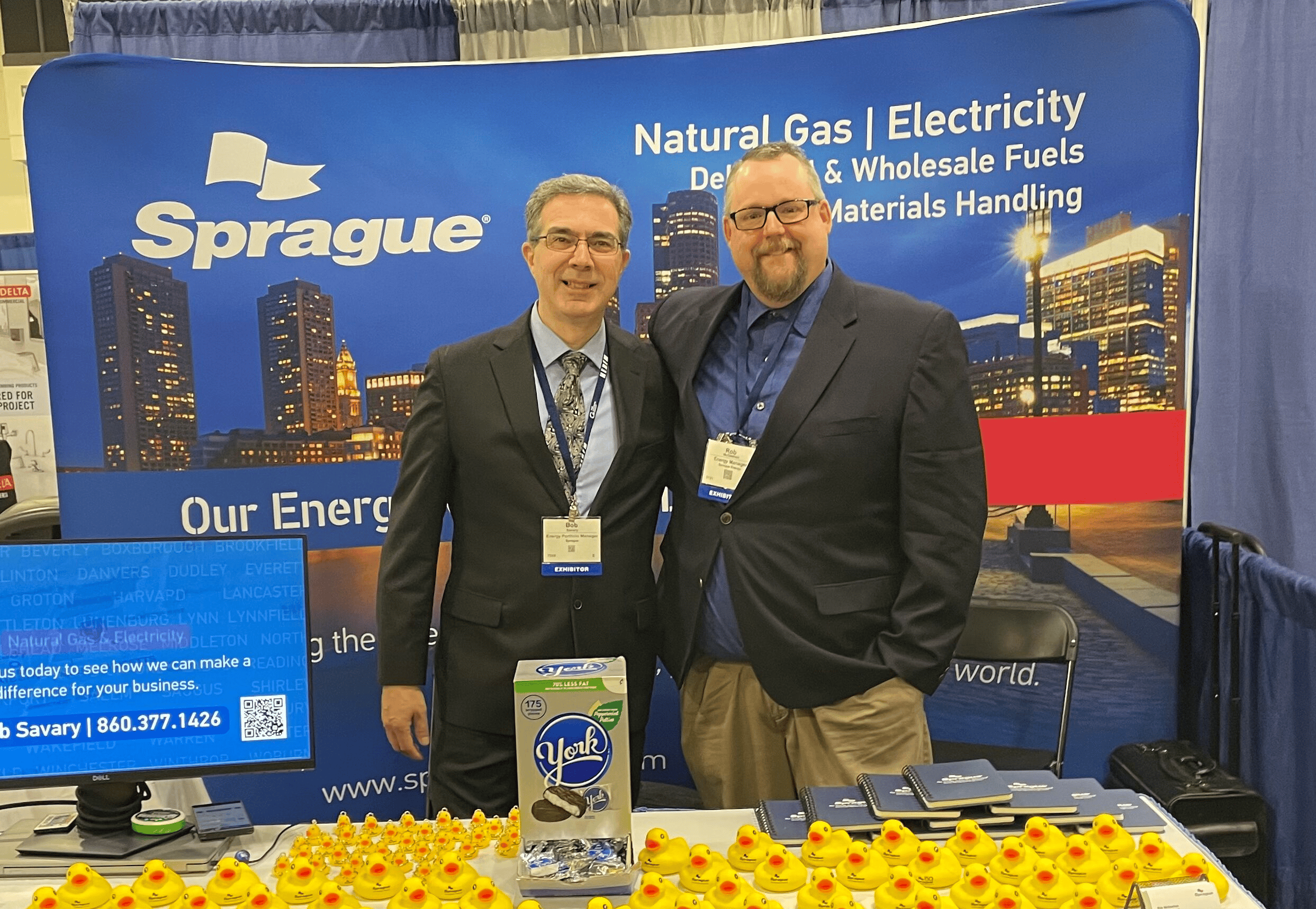 Two Sprague Energy representatives at a trade show booth promoting natural gas, electricity, and fuel services, with branded materials and rubber duck giveaways on display.