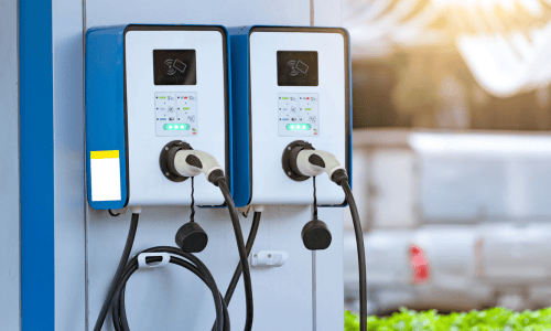 Two electric vehicle (EV) charging stations with plugged-in cables are shown in an outdoor setting, with blurred vehicles and greenery in the background.