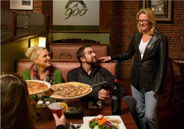 Group dining scene at a cozy Italian restaurant, with four people seated around a wooden table enjoying two pizzas, a salad, wine, and beer, while another person stands nearby. The background features a rustic brick wall and signage reading “900° Simply Italian Pizza.”