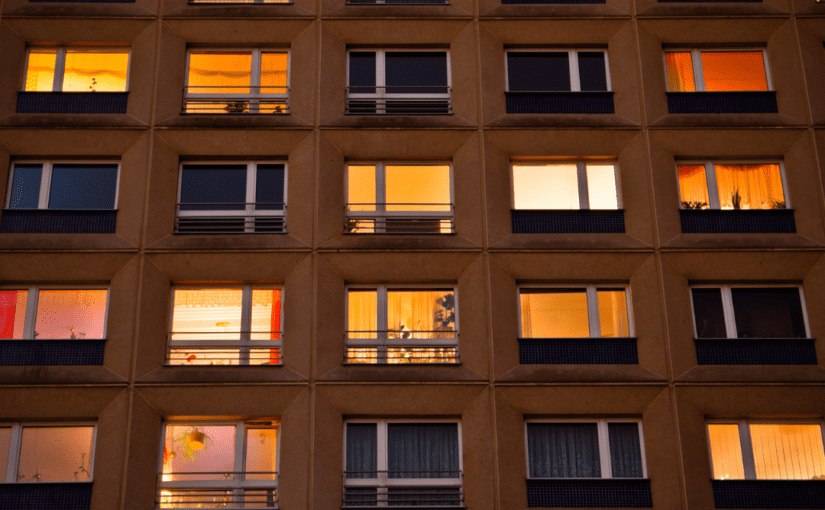 Exterior view of a multi-story apartment building at dusk, with warm lights glowing from the windows of various units, creating a grid-like pattern across the facade.